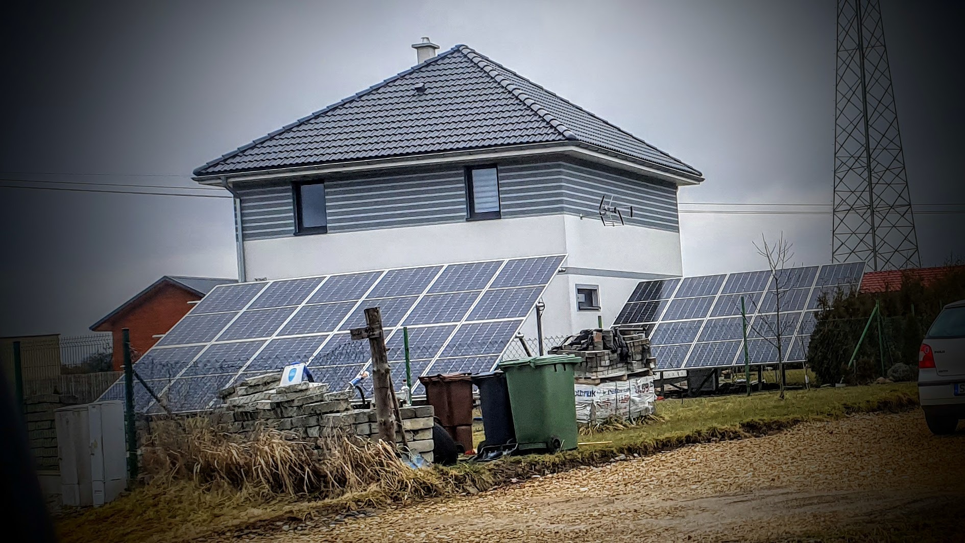 Panele słoneczne zamontowane na ziemi przed nowoczesnym domem z szarą elewacją i ciemnym dachem, widoczne śmietniki i sterta kostki brukowej, pochmurne niebo w tle.
