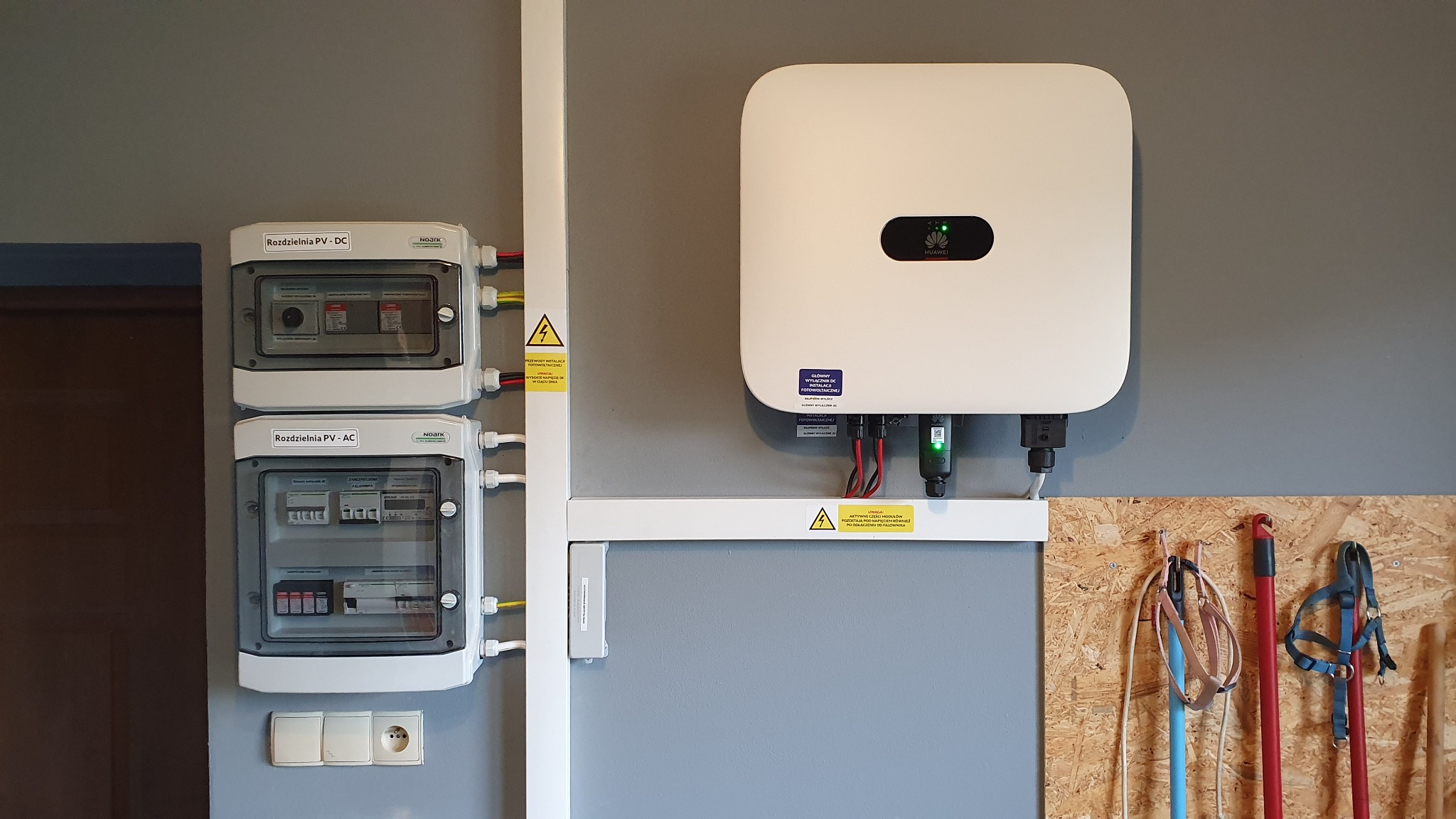 Rozdzielnice PV-DC i PV-AC, inwerter Huawei na szarej ścianie. Widoczne kable, gniazdko i narzędzia na ścianie z płyty OSB. Instalacja fotowoltaiczna w garażu.