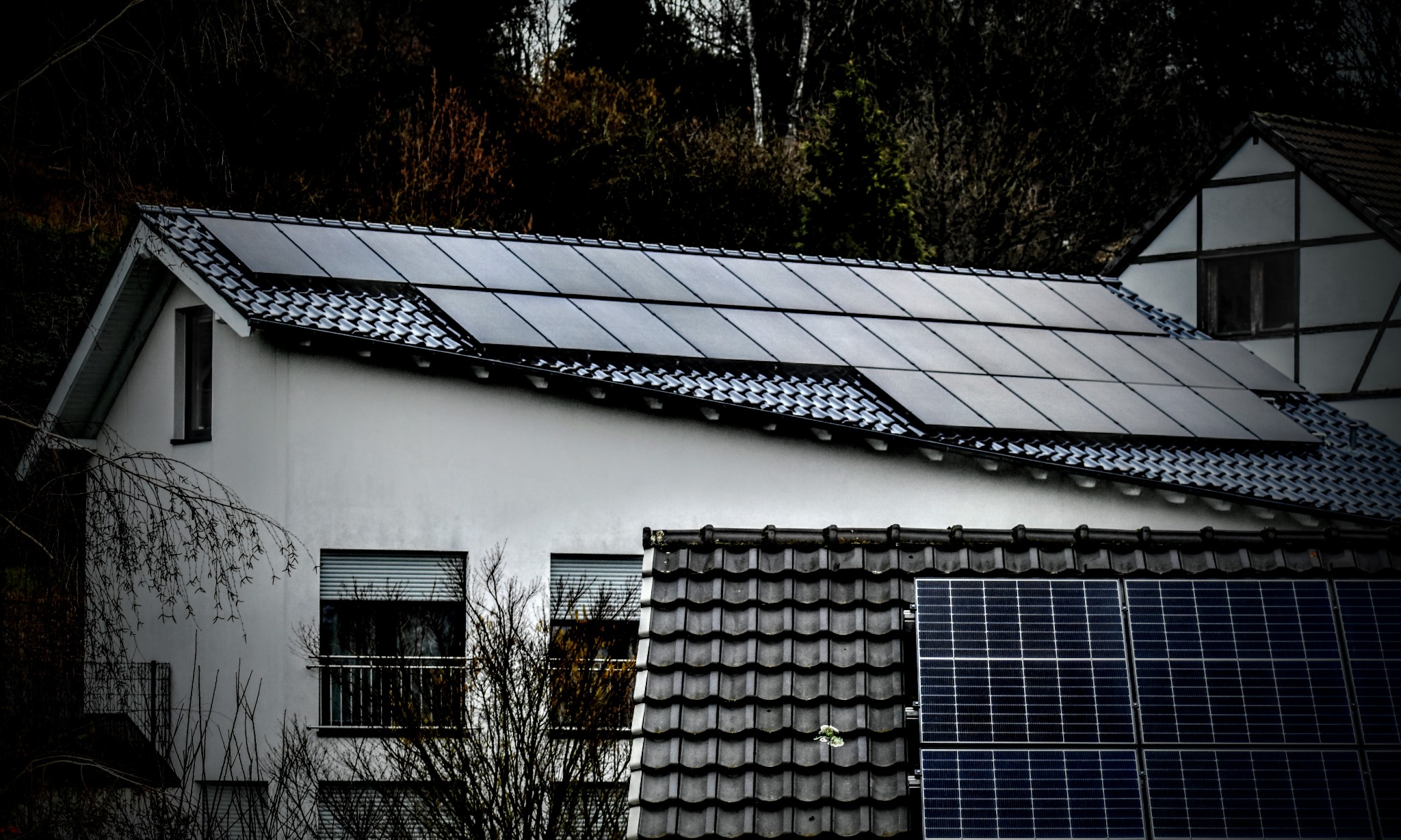 Dom z panelami fotowoltaicznymi na dachu, widoczne panele na dwóch poziomach dachu, tło z drzewami i fragmentem drugiego budynku.