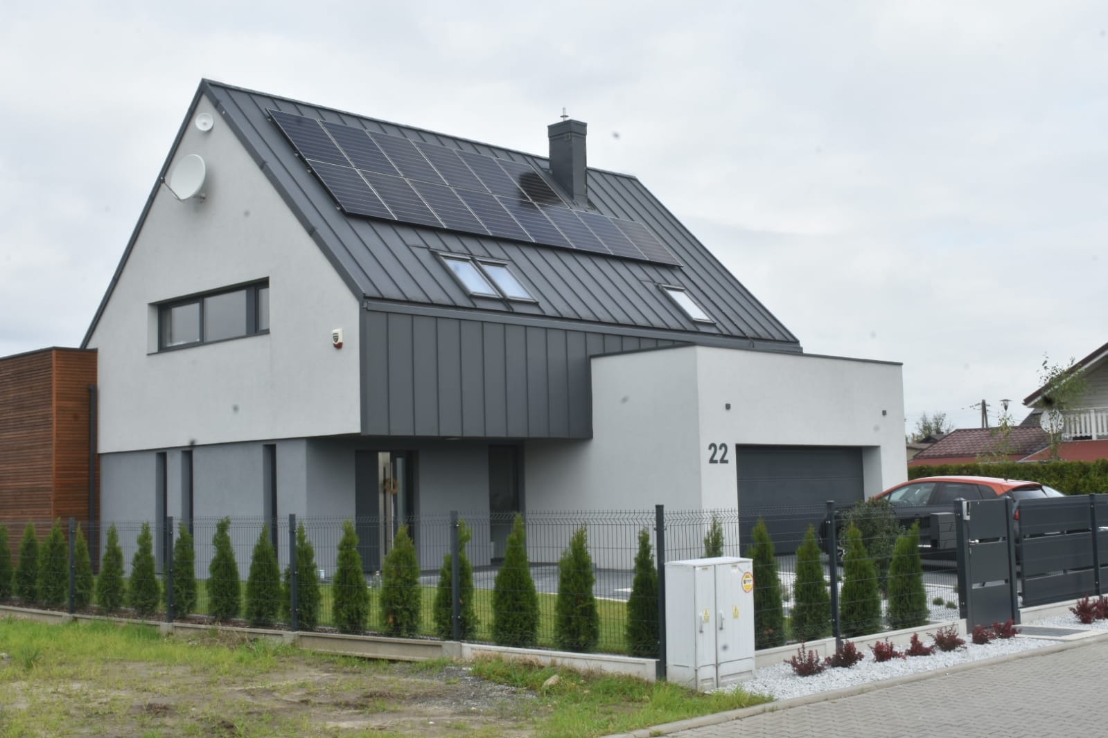 Nowoczesny dom jednorodzinny z panelami fotowoltaicznymi na grafitowym dachu. Biała elewacja, numer domu 22, szara brama i ogrodzenie. W tle pochmurne niebo.