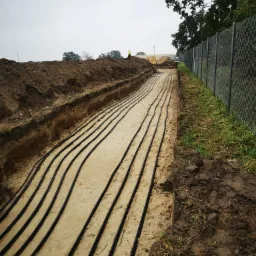 Wykop z ułożonymi rzędami czarnych rur w piasku, widoczni pracownicy w oddali i ogrodzenie z siatki.