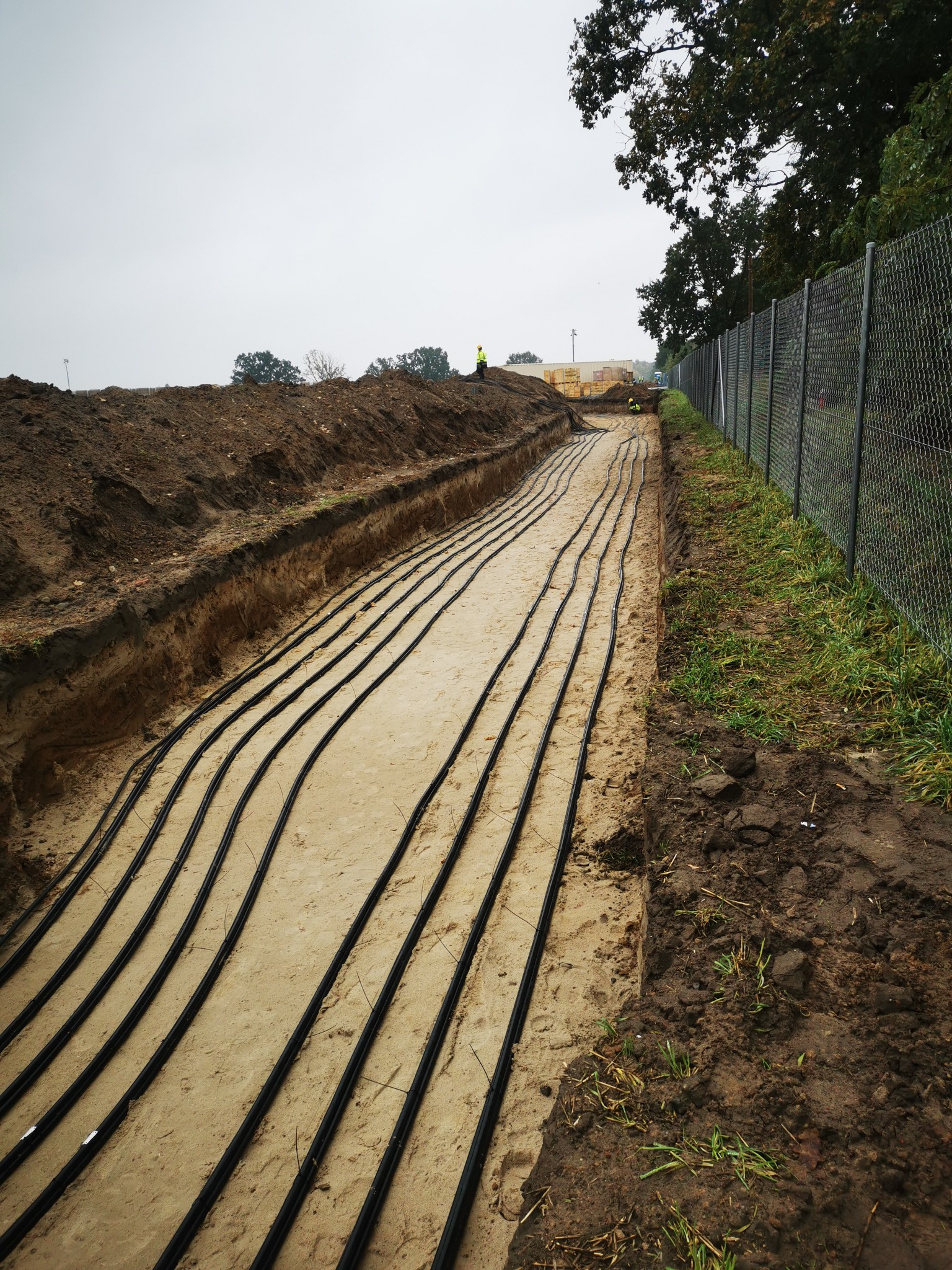 Wykop z ułożonymi rzędami czarnych rur w piasku, widoczni pracownicy w oddali i ogrodzenie z siatki.