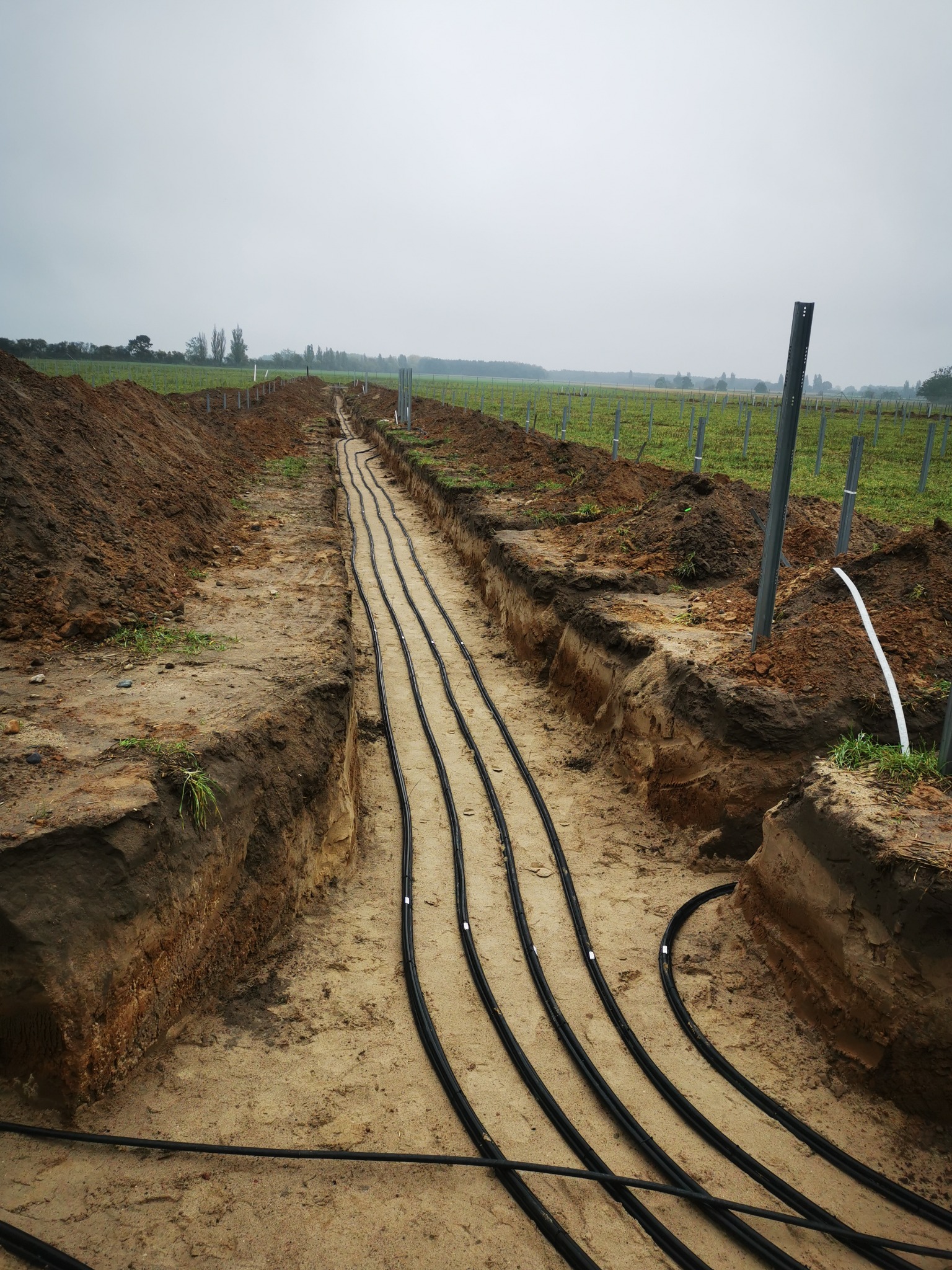 Wykop z ułożonymi rurami instalacji w Olsztynie, widok z góry na cztery czarne rury w piaskowym wykopie, w tle pola i słupy.