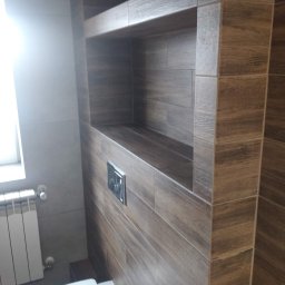 HOMME BUD Krzysiek - Nowoczesna łazienka z WC i zabudowaną półką wyłożoną brązowymi płytkami imitującymi drewno, widoczny fragment grzejnika i okna.