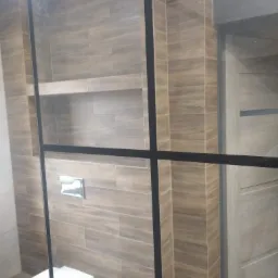 Nowoczesna łazienka z drewnopodobnymi płytkami ułożonymi poziomo, zabudowaną półką, toaletą wiszącą i kabiną prysznicową z czarnymi profilami.