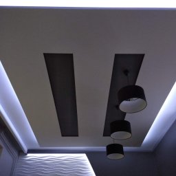 HOMME BUD Krzysiek - Nowoczesny sufit podwieszany z oświetleniem LED wzdłuż krawędzi i czarnymi lampami wiszącymi na tle dekoracyjnej ściany z falistym wzorem.