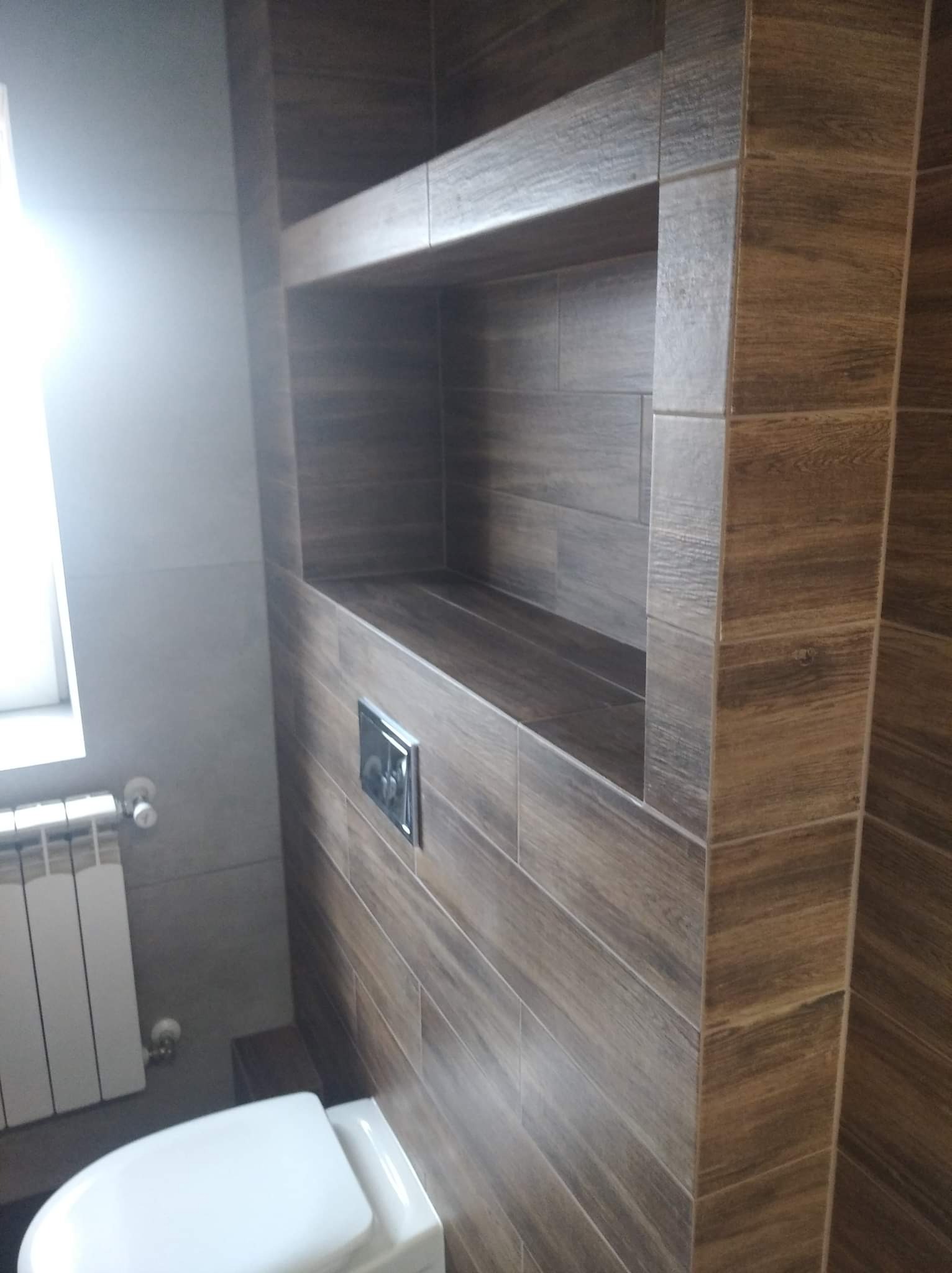 Nowoczesna łazienka z WC i zabudowaną półką wyłożoną brązowymi płytkami imitującymi drewno, widoczny fragment grzejnika i okna.