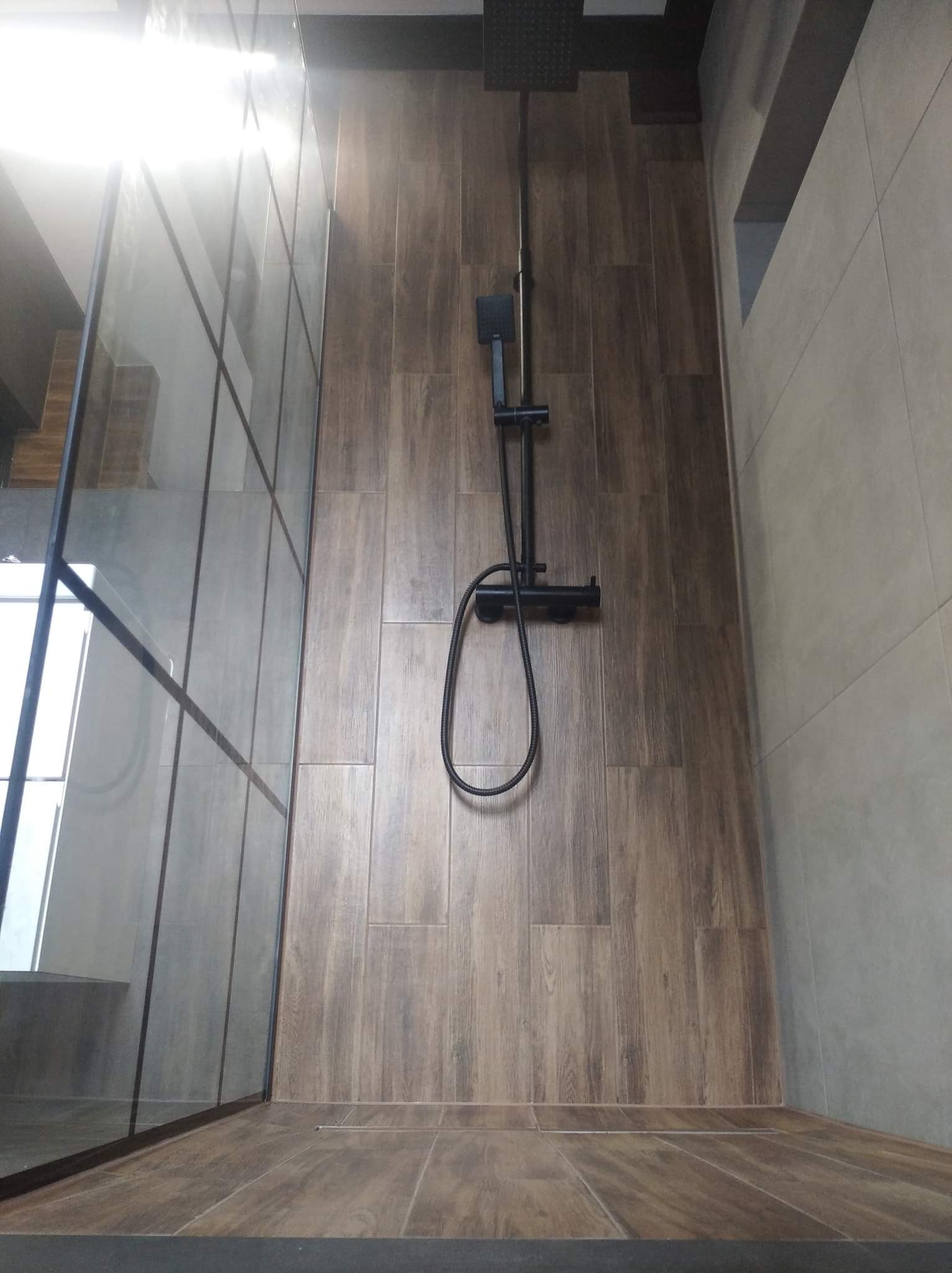 Nowoczesna kabina prysznicowa typu walk-in z czarną armaturą na tle płytek imitujących drewno i szarych płytek ściennych.