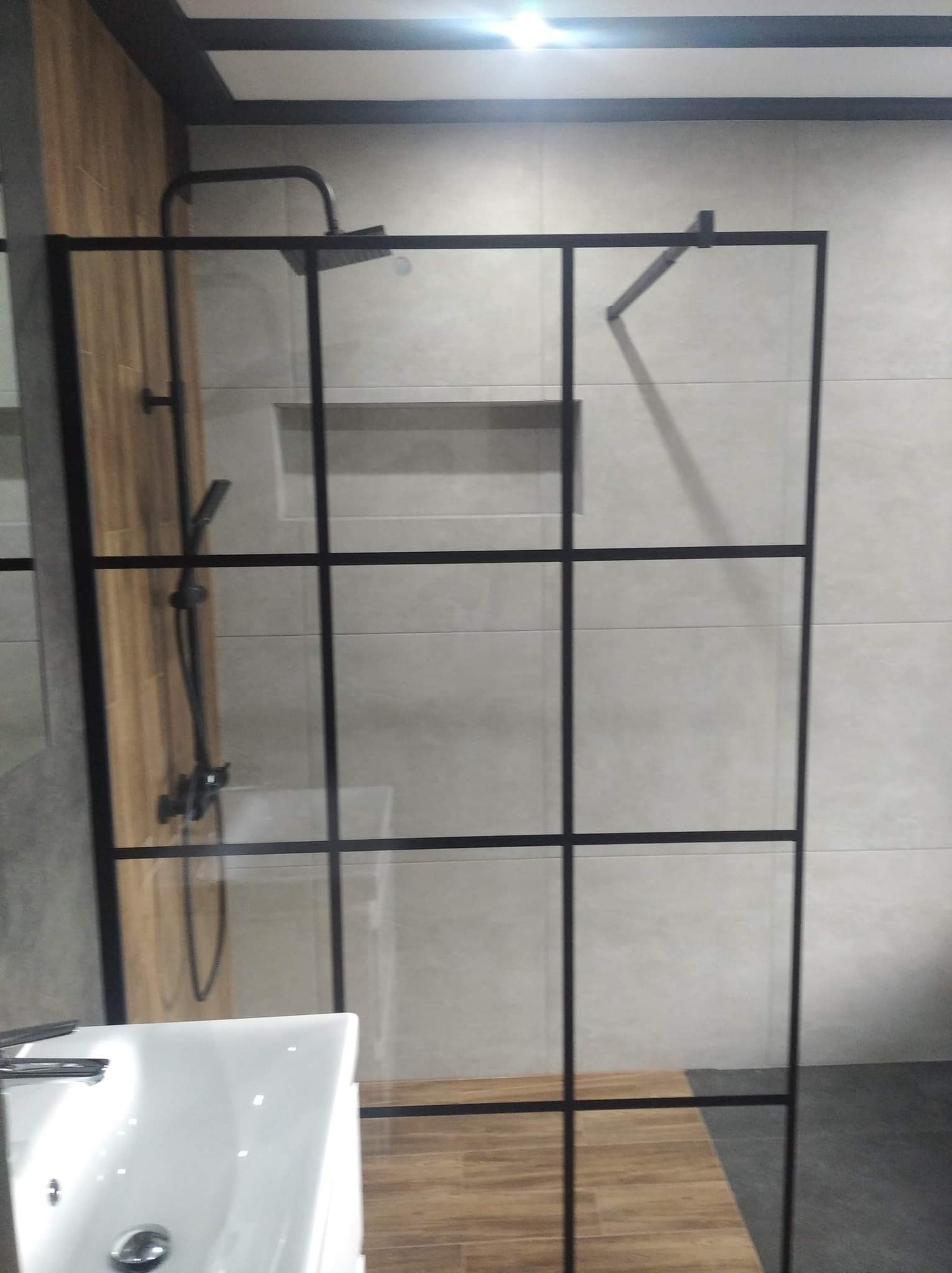 Kabina prysznicowa z czarnymi profilami w nowoczesnej łazience, widoczna armatura i umywalka.