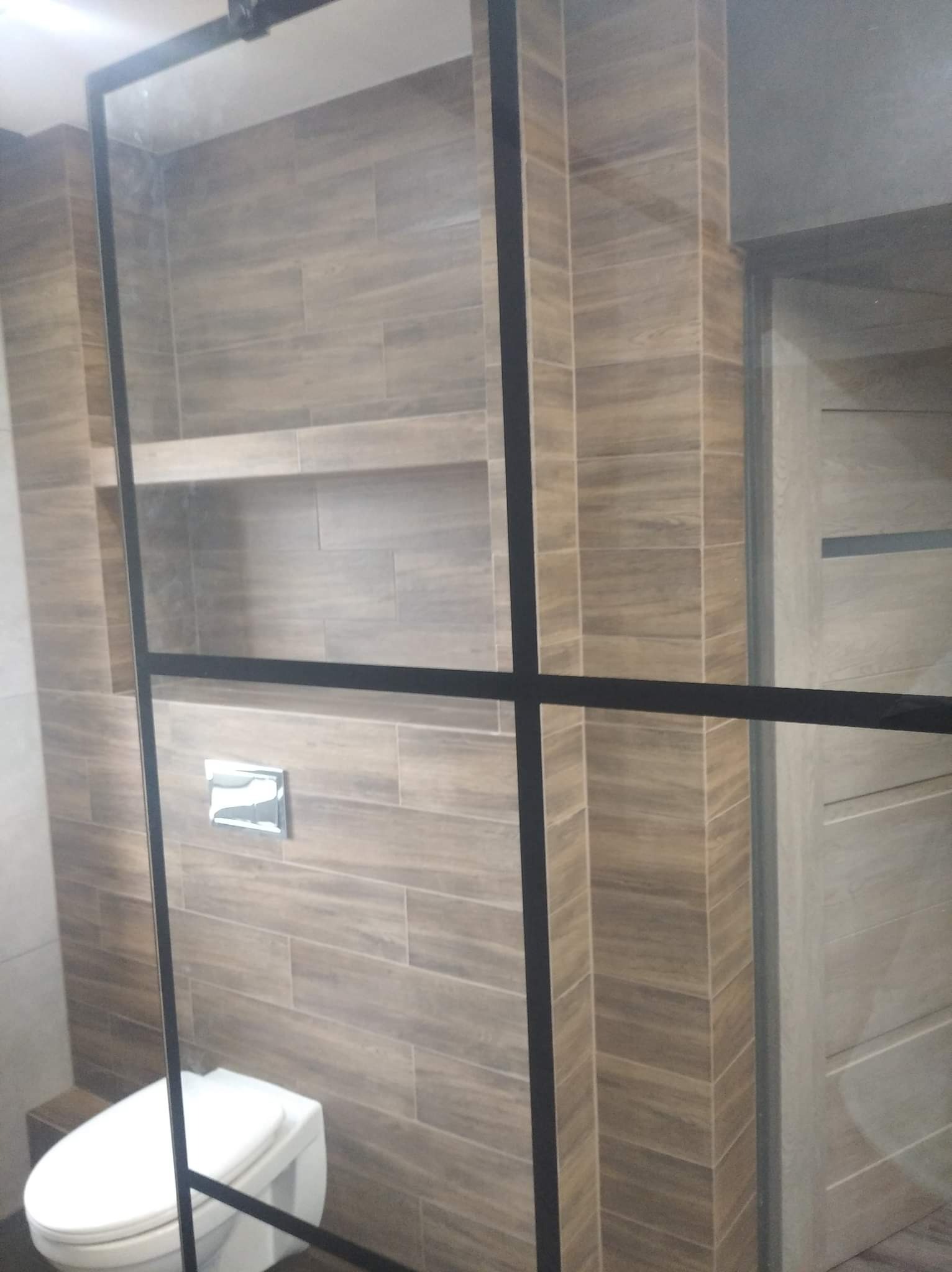 Nowoczesna łazienka z drewnopodobnymi płytkami ułożonymi poziomo, zabudowaną półką, toaletą wiszącą i kabiną prysznicową z czarnymi profilami.