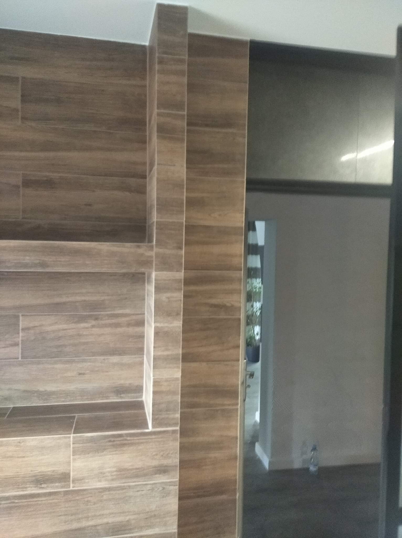 Ściana z płytek imitujących drewno z wnęką, widok na sąsiednie pomieszczenie przez otwarte drzwi.