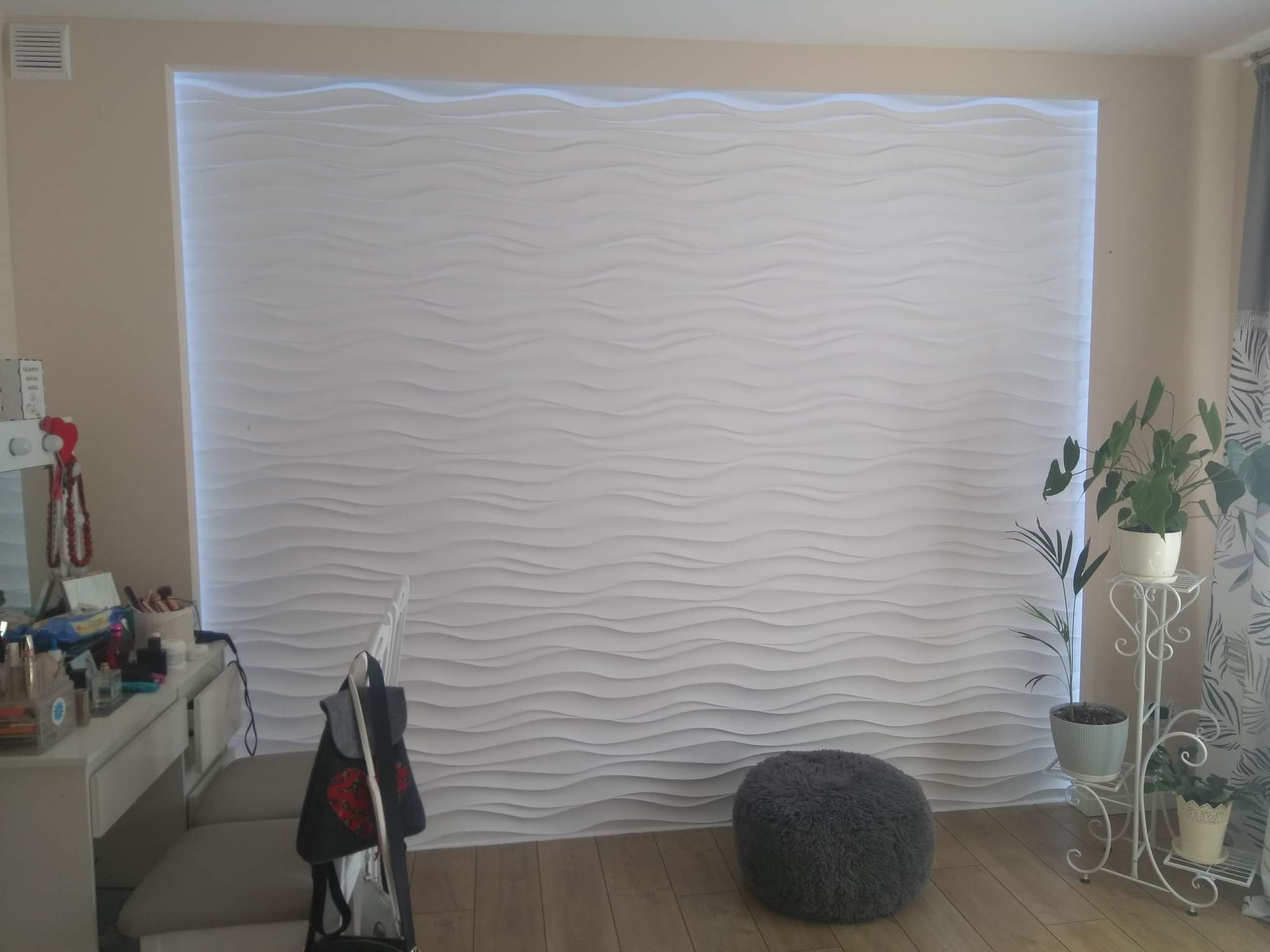 Biała, trójwymiarowa ściana dekoracyjna z subtelnym podświetleniem LED, widoczna w tle pokoju z kremowymi ścianami, drewnianą podłogą, toaletką z lustrem po lewej i kwietnikiem z roślinami po prawej.