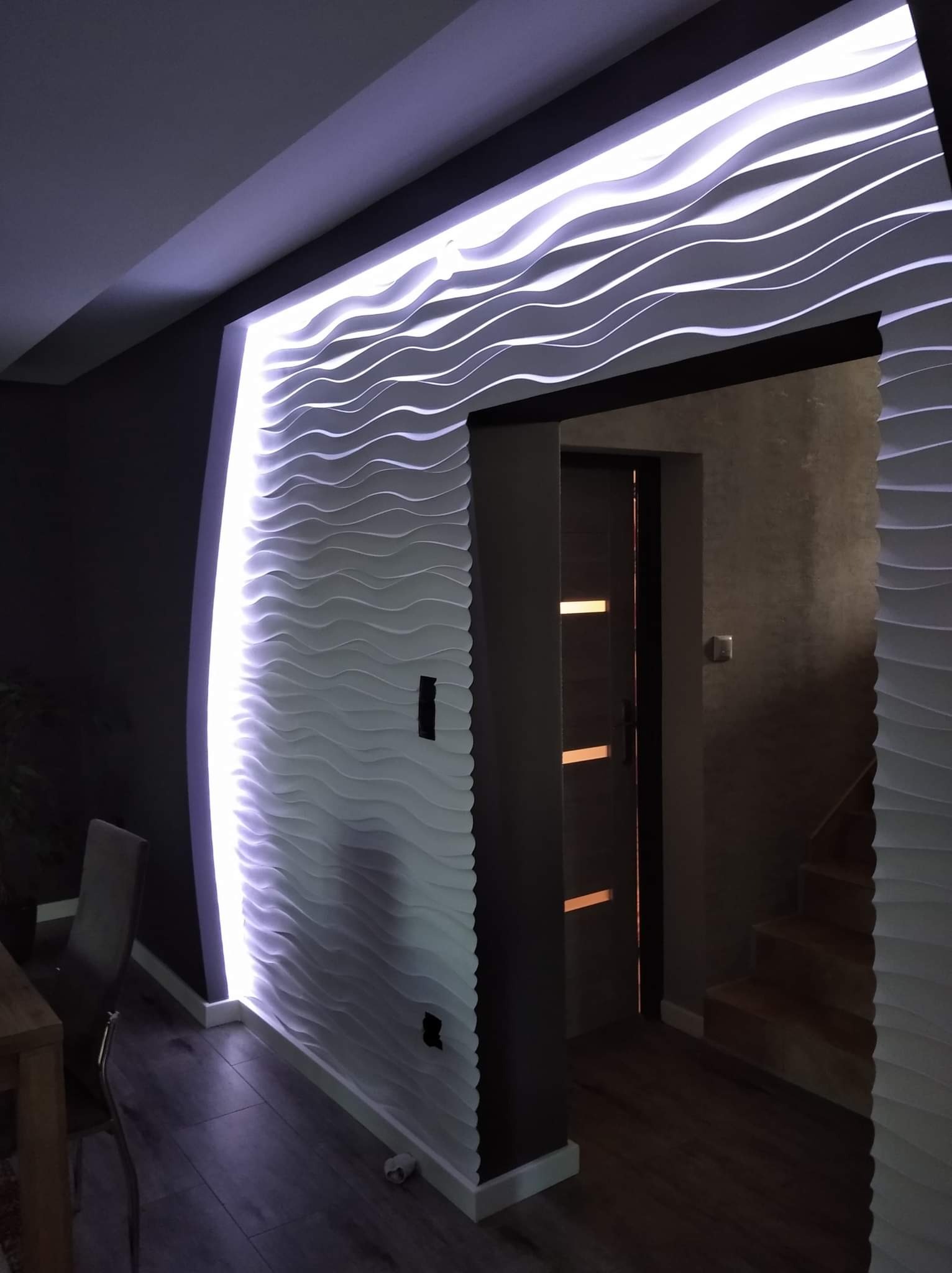 Nowoczesna ściana z paneli 3D o falistym wzorze, podświetlona fioletowym światłem LED, tworząca dekoracyjny element wnętrza obok wejścia do pomieszczenia.