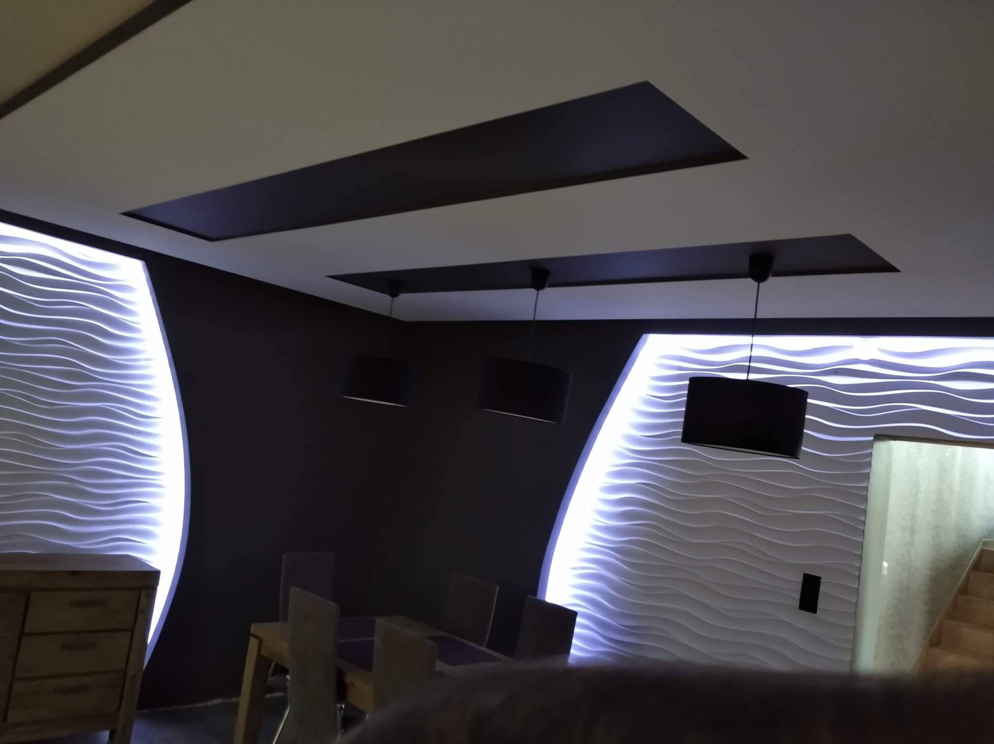 Nowoczesne wnętrze jadalni z podświetlanymi panelami ściennymi o falistej strukturze, czarnymi elementami sufitu i minimalistycznymi lampami wiszącymi.