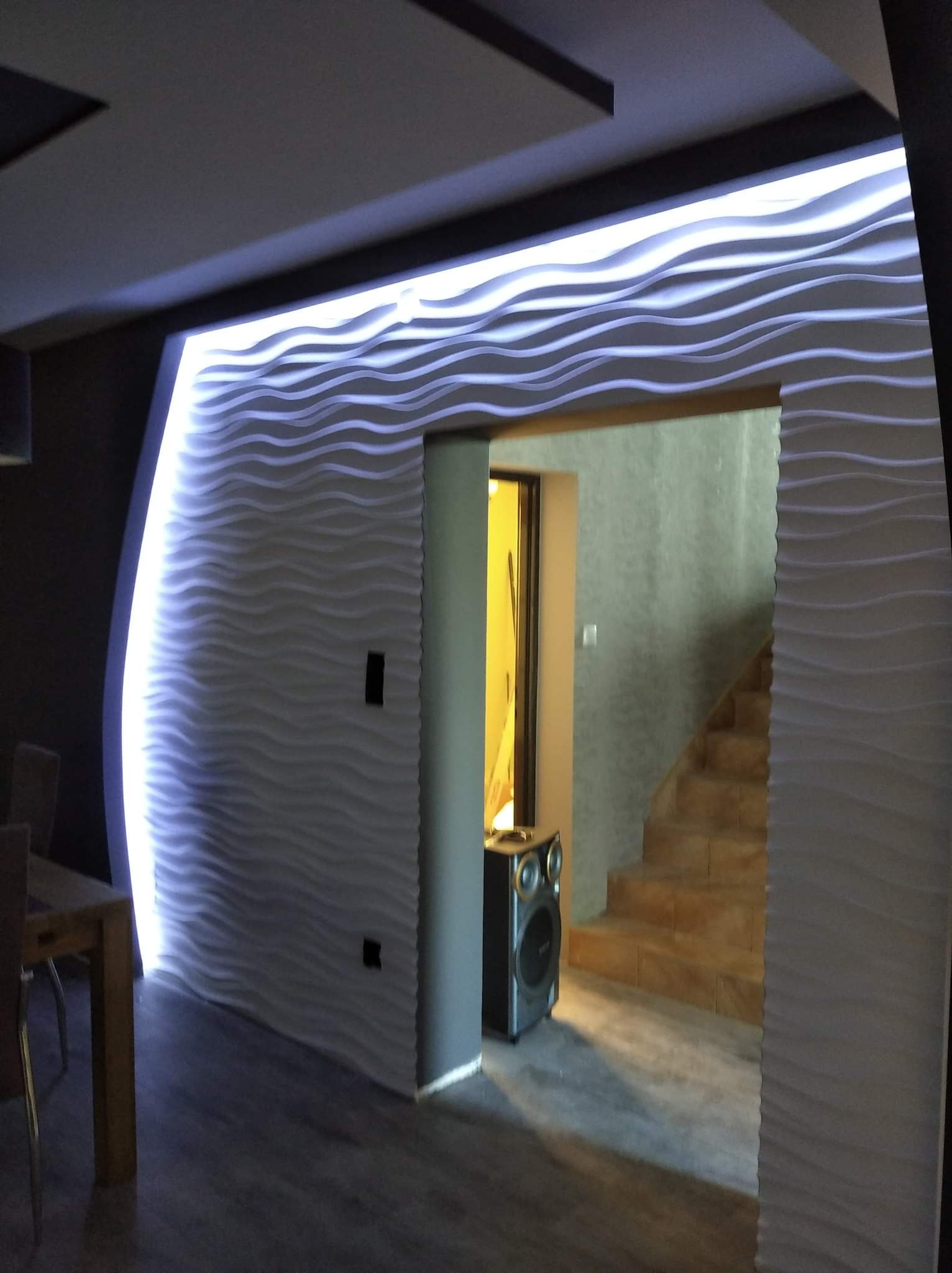 Nowoczesne wnętrze z dekoracyjną ścianą 3D o falistym wzorze, podświetloną taśmą LED, widok na schody i głośnik w przejściu.