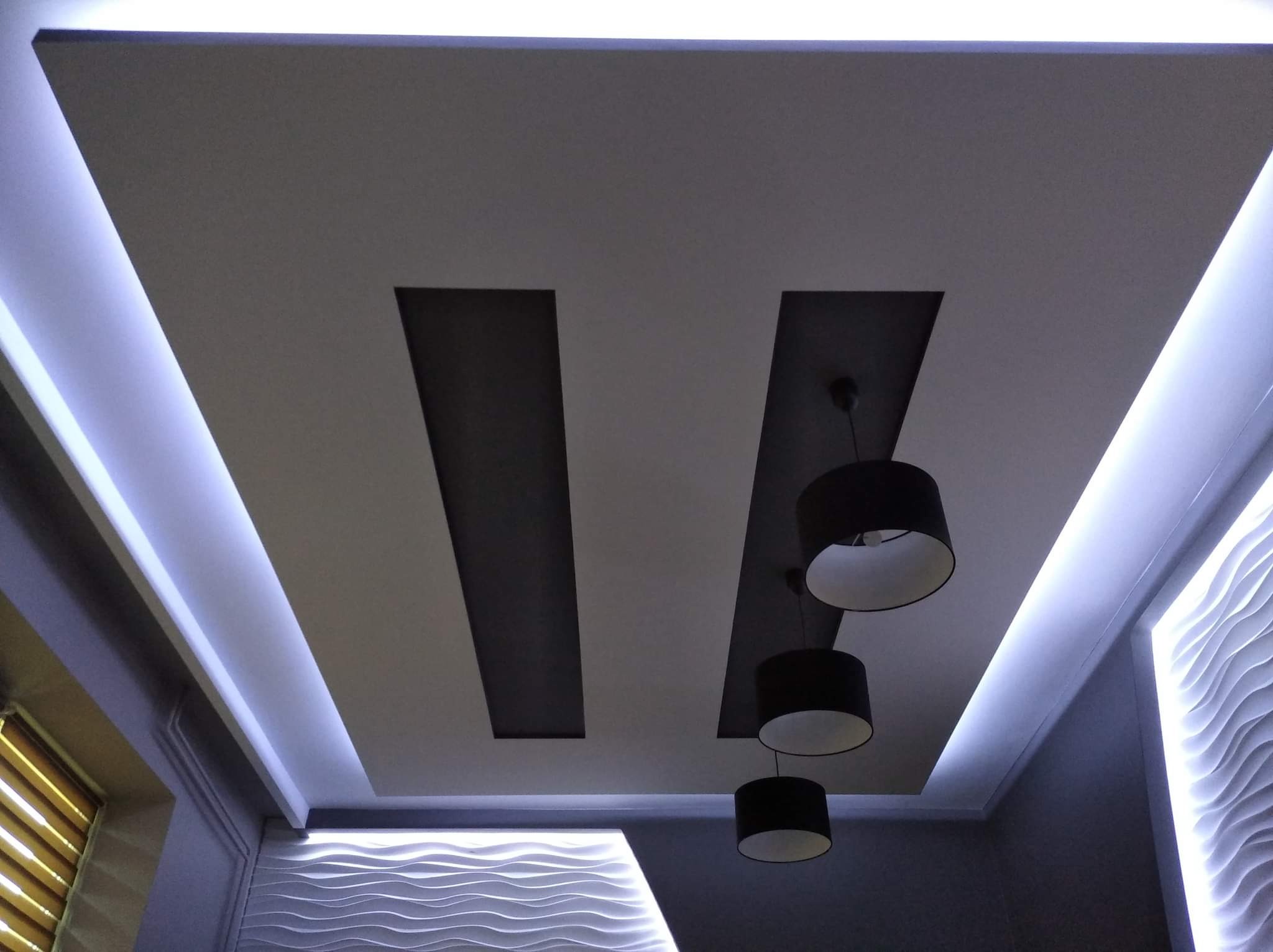 Nowoczesny sufit podwieszany z oświetleniem LED wzdłuż krawędzi i czarnymi lampami wiszącymi na tle dekoracyjnej ściany z falistym wzorem.