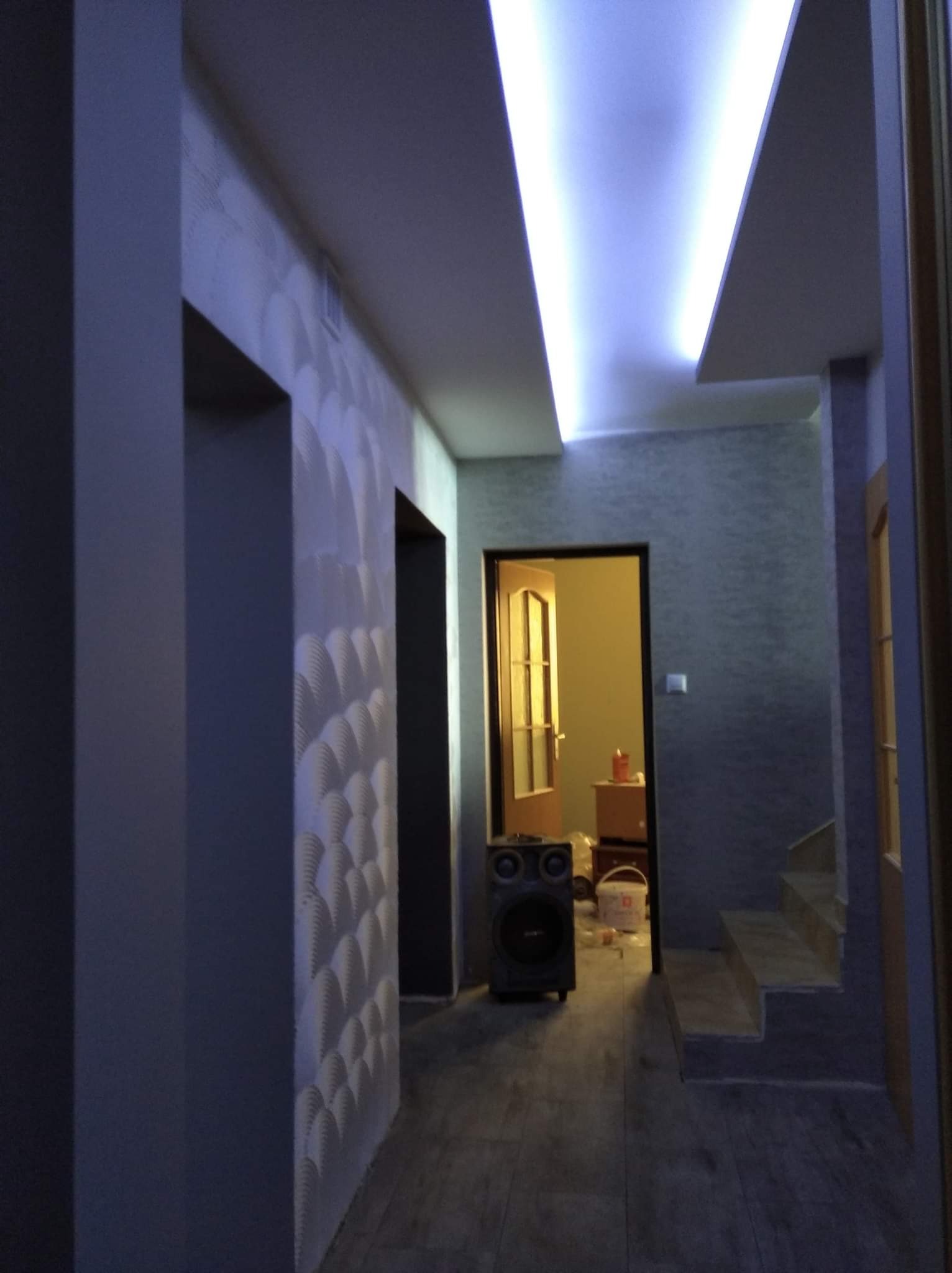 Korytarz po remoncie z oświetleniem LED na suficie, dekoracyjną ścianą z wzorem 3D i widocznymi schodami oraz sprzętem audio w tle.