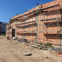 Wznoszona ściana z czerwonej cegły, z widocznymi rusztowaniami i przygotowanym terenem budowy, w tle budynek mieszkalny.