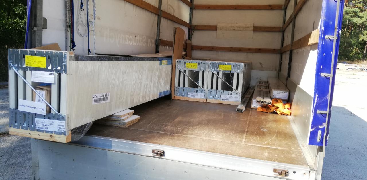 Wnętrze przestrzeni ładunkowej busa z zapakowanymi i zabezpieczonymi elementami konstrukcyjnymi, skrzynie z metalowymi wzmocnieniami, widoczne etykiety i ostrzeżenia.