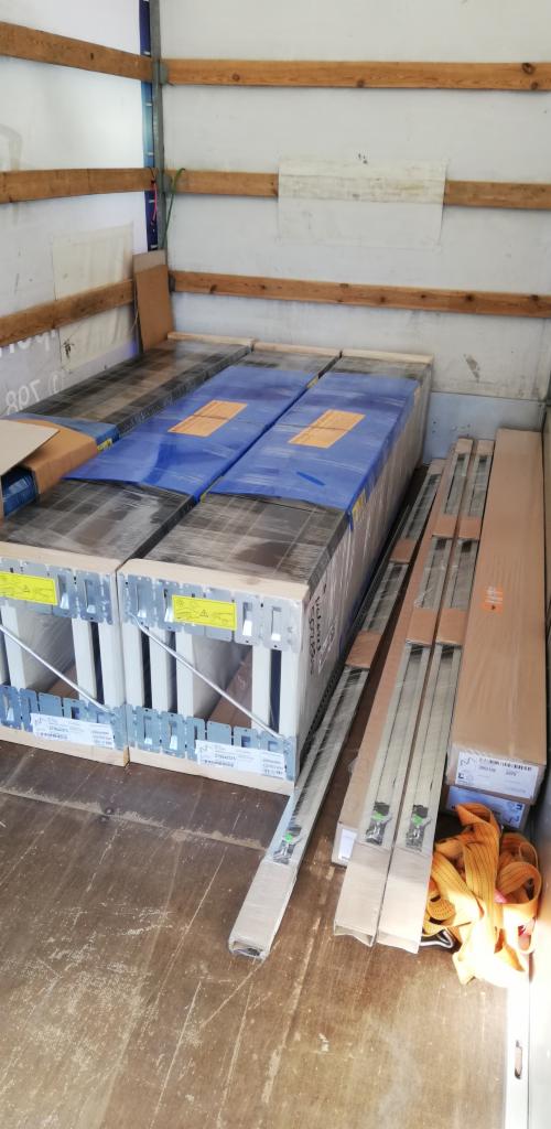 Wnętrze busa załadowane paletami z oknami w metalowych ramach i niebieską folią, oraz długimi kartonami z roletami, zabezpieczone żółtymi pasami transportowymi.