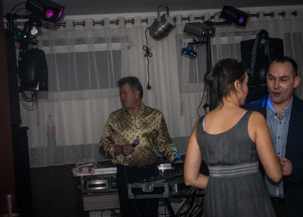 Występujący mężczyzna z mikrofonem w złotej koszuli, mikser, oświetlenie sceniczne i para na parkiecie podczas imprezy z muzyką na żywo.