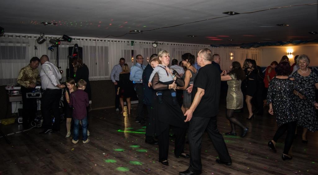 Roztańczona sala balowa z parkietem, oświetleniem dyskotekowym i DJ-em w tle. W centrum kadru goście weselni w ruchu, w tym kobieta trzymająca dziecko.