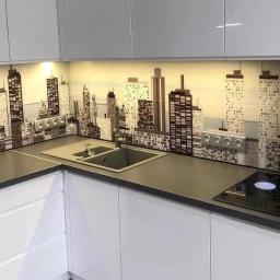 Polgraf - Nowoczesna kuchnia z białymi szafkami i szarym blatem, ozdobiona szklanym panelem ściennym z motywem panoramy miasta w odcieniach szarości, z widocznymi gniazdkami elektrycznymi.