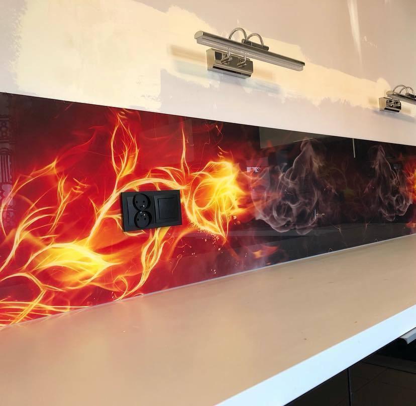 Szklany panel ścienny z motywem ognia i dymu nad blatem, czarne gniazdko elektryczne na tle płomieni, oświetlenie halogenowe zamontowane nad panelem.