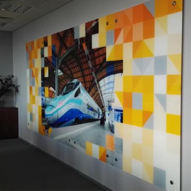 Duży panel dekoracyjny z motywem pociągu w hali dworcowej, wkomponowany w geometryczny wzór z żółtych, pomarańczowych i szarych kwadratów, zamocowany na ścianie w biurze.