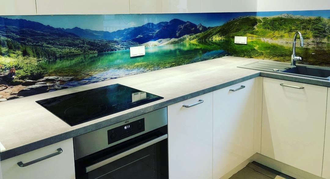 Szklany panel ścienny z motywem górskiego jeziora nad blatem kuchennym z płytą indukcyjną i piekarnikiem AEG w nowoczesnej białej kuchni.