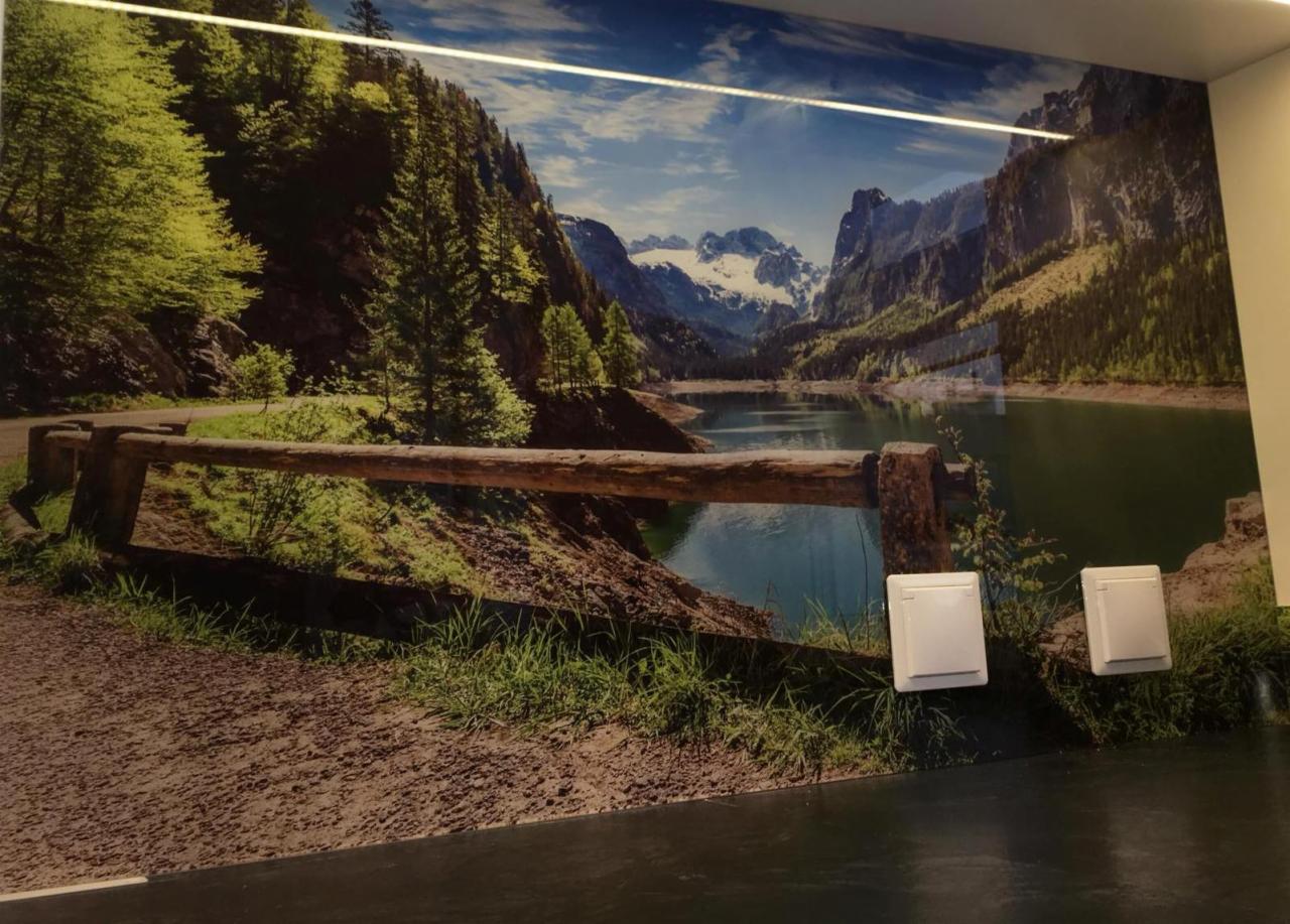 Ściana biura oklejona fototapetą z widokiem na górskie jezioro i drewnianą barierkę, z dwoma białymi elementami instalacji elektrycznej na pierwszym planie.