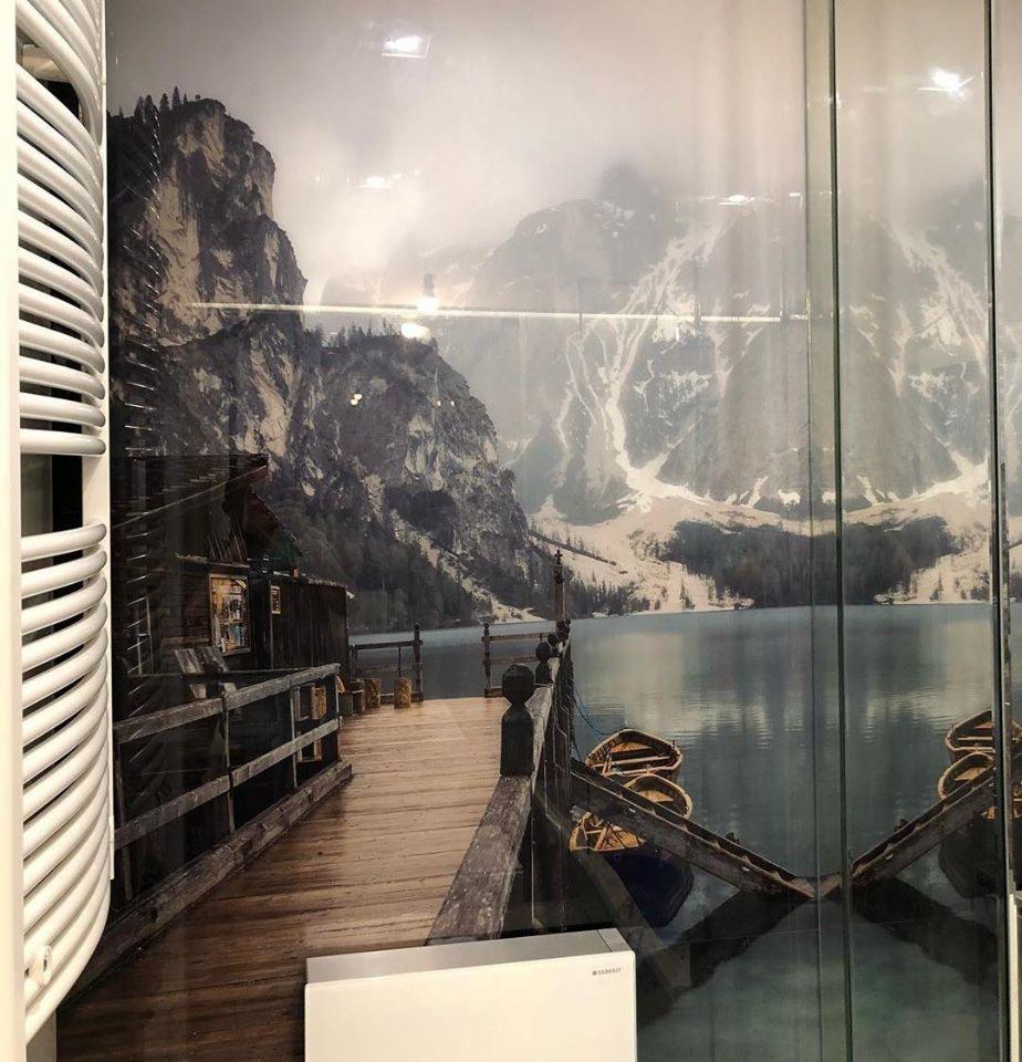 Wielkoformatowy wydruk na ścianie przedstawiający drewniane molo nad jeziorem z łodziami na tle górskiego krajobrazu, widoczny grzejnik i fragment szklanej ściany.
