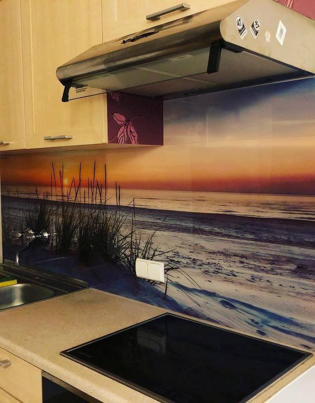 Szklany panel ścienny w kuchni z motywem plaży o zachodzie słońca, imitujący widok na morze z trawami, zamontowany nad płytą indukcyjną i zlewem.