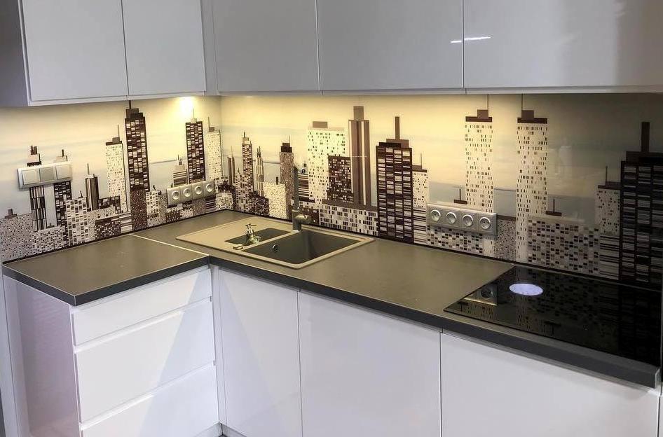 Nowoczesna kuchnia z białymi szafkami i szarym blatem, ozdobiona szklanym panelem ściennym z motywem panoramy miasta w odcieniach szarości, z widocznymi gniazdkami elektrycznymi.