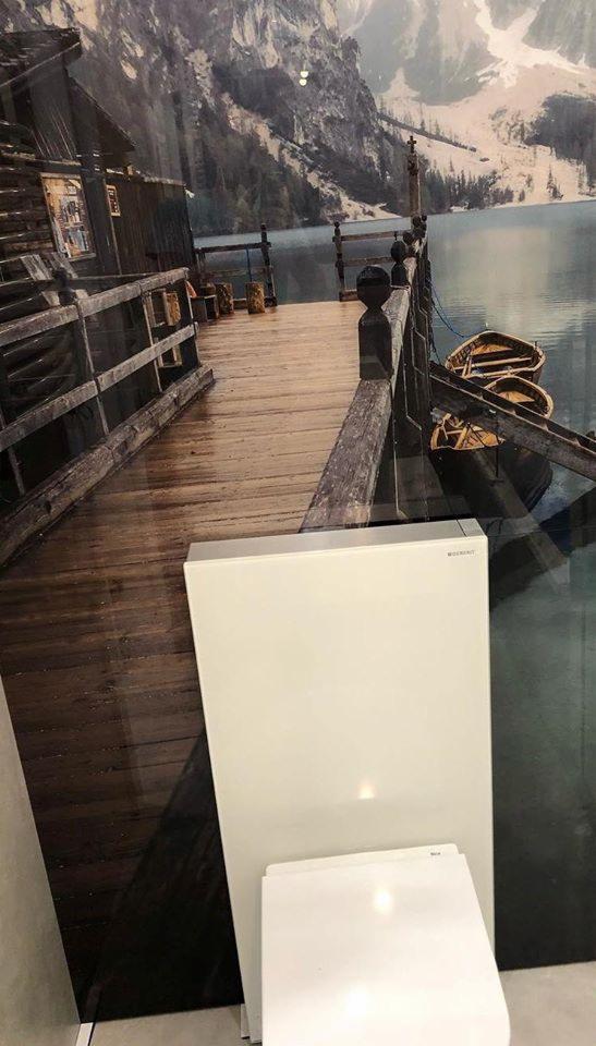 Biały kompaktowy zestaw toaletowy na tle fototapety przedstawiającej drewniany pomost nad jeziorem z łodziami i górskim krajobrazem.