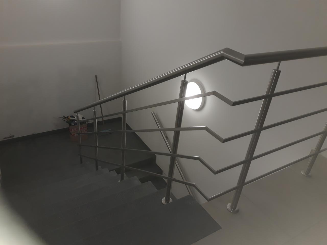 Wewnętrzna balustrada nierdzewna ze schodami, z geometrycznym wzorem, oświetlona okrągłą lampą na ścianie, widok z dołu.