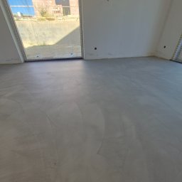 Usługi budowlane - Szara, gładka powierzchnia w pomieszczeniu z dużym oknem. Wykończona podłoga w minimalistycznym stylu. Widoczne ślady po pracy na powierzchni.