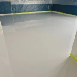 Biała, gładka powierzchnia podłogi po wylaniu, zabezpieczona folią malarską przy ścianach. Niebieska ściana w tle, przygotowana do dalszych prac wykończeniowych.