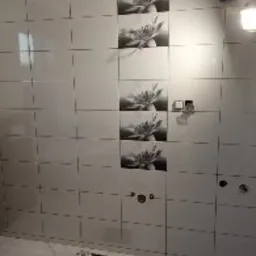 Ściana łazienki wyłożona białymi, błyszczącymi płytkami z motywem kwiatowym w pionowym pasie, widoczne otwory pod armaturę i gniazdka elektryczne, podłoga z jasnego marmuru, niedokończona realizacja.