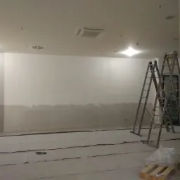 Przygotowane do malowania wnętrze biura z dwoma rozstawionymi drabinami, zabezpieczoną podłogą i częściowo wykończonymi ścianami.