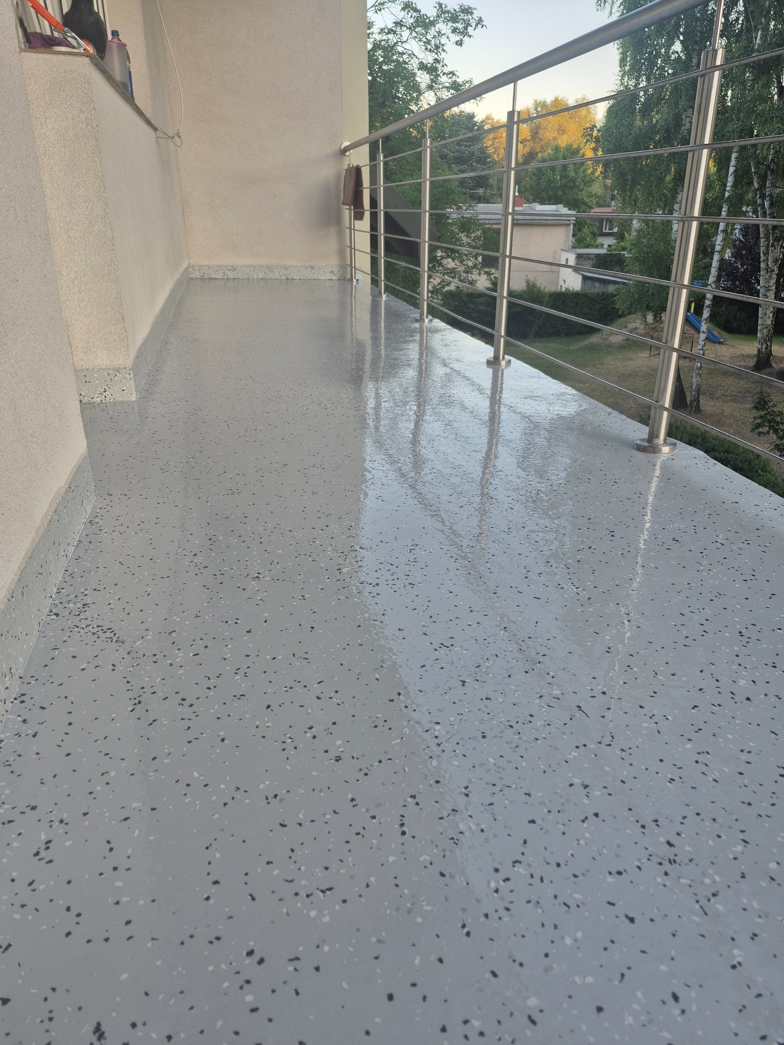 Balkon z szarą, błyszczącą posadzką z drobnymi, ciemnymi plamkami. Metalowa balustrada i widok na zieleń w tle. Gładka powierzchnia odbija światło.