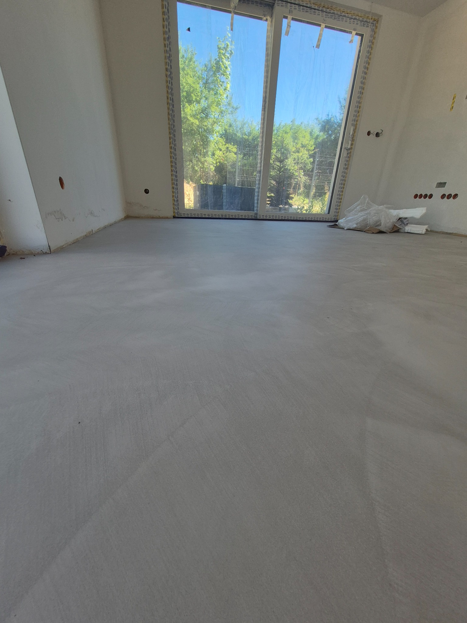 Szara, gładka posadzka w jasnym pokoju z dużym oknem balkonowym. Widoczne ślady po szpachlowaniu na ścianach. W tle zieleń drzew za oknem.