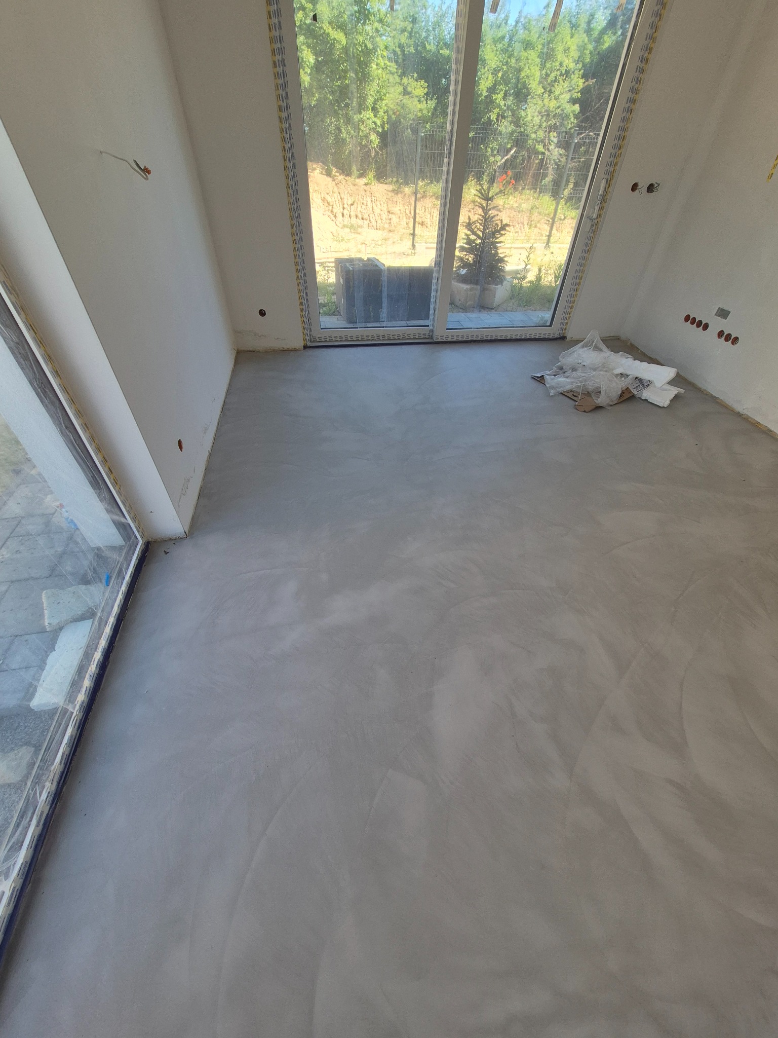 Jasnoszara, gładka powierzchnia w pomieszczeniu z dużym oknem tarasowym. Widoczne ślady po aplikacji i delikatny połysk. W tle zieleń ogrodu.
