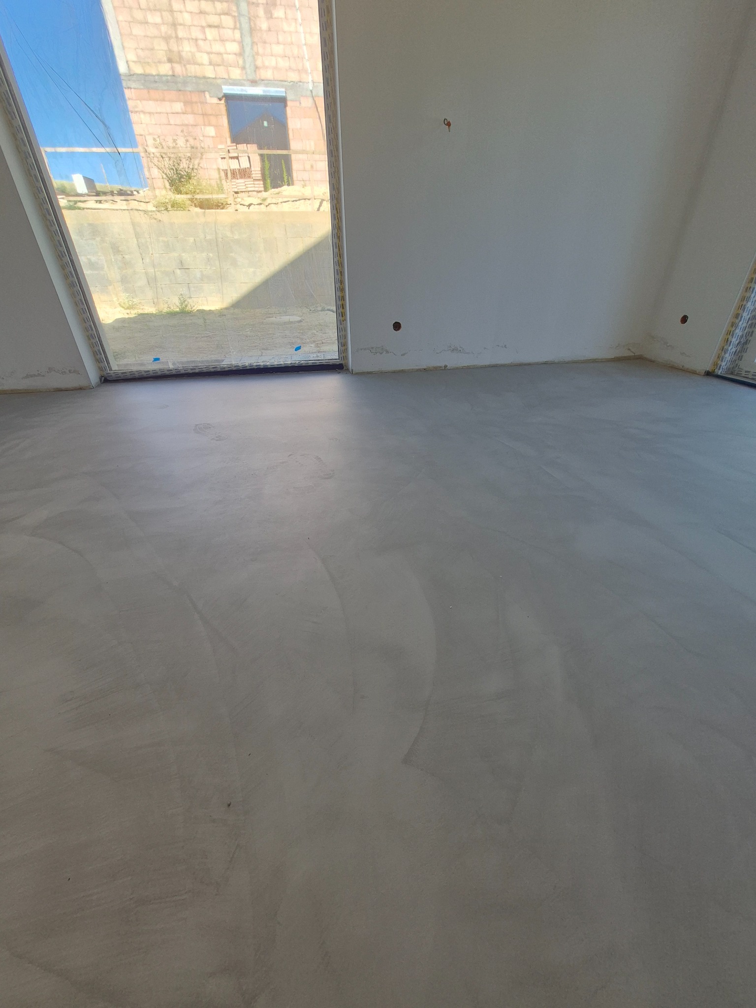 Szara, gładka powierzchnia w pomieszczeniu z dużym oknem. Wykończona podłoga w minimalistycznym stylu. Widoczne ślady po pracy na powierzchni.