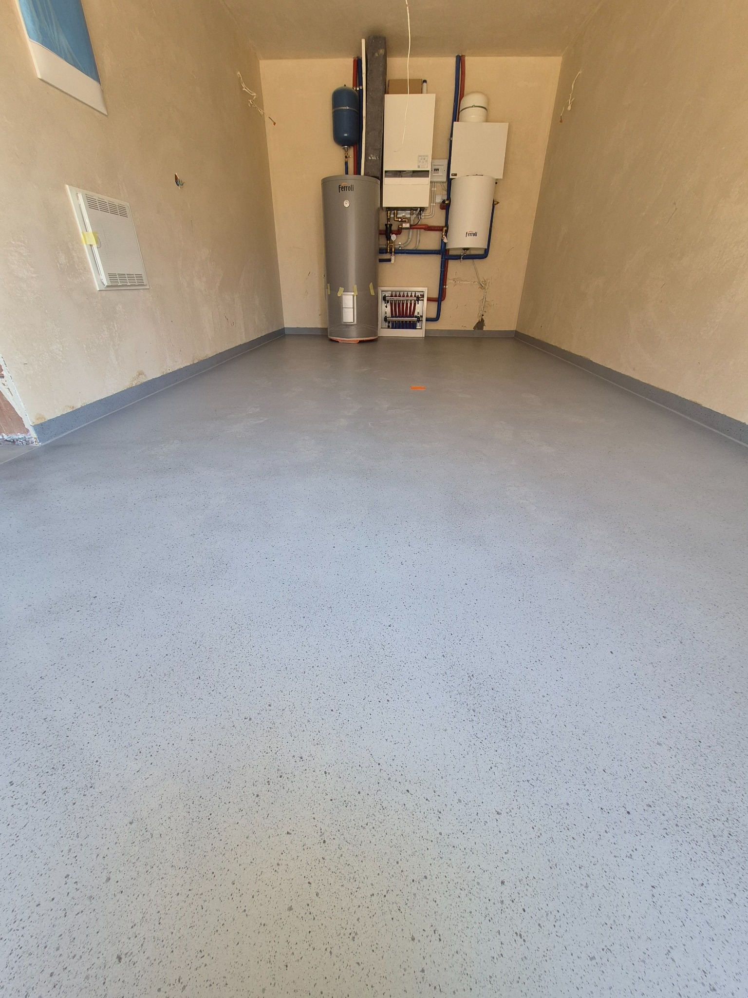 Jasnoszara, gładka powierzchnia podłogi w pomieszczeniu z instalacją grzewczą na ścianie. Widoczny detal wykończenia przy ścianie i delikatna faktura posadzki.