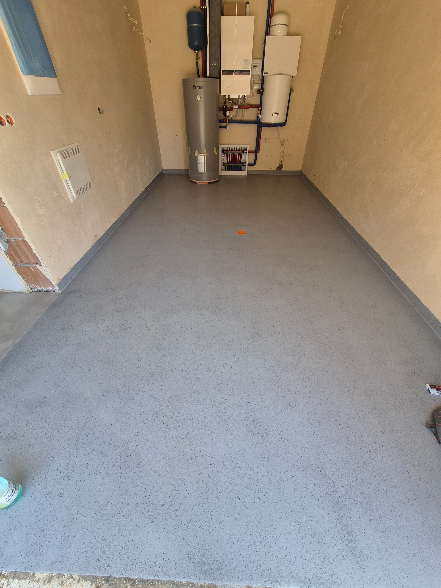 Szara posadzka żywiczna w pomieszczeniu technicznym z piecem i bojlerem. Nowa, gładka powierzchnia z delikatnym wzorem. Widoczne elementy instalacji grzewczej.