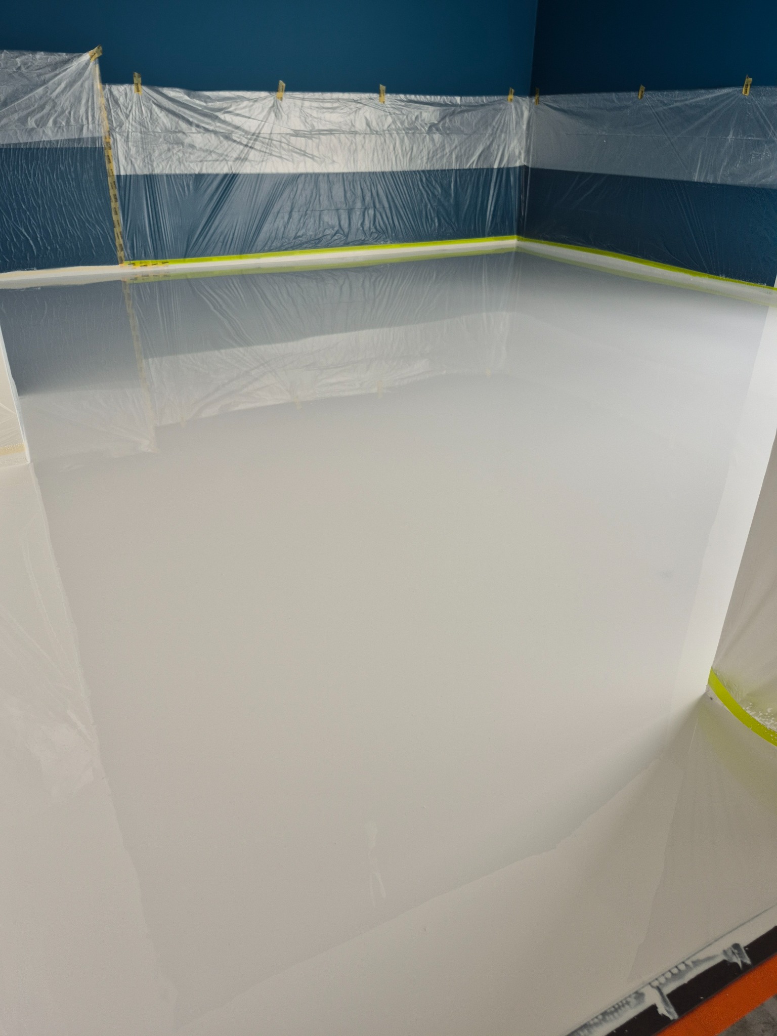 Biała, gładka powierzchnia podłogi po wylaniu, zabezpieczona folią malarską przy ścianach. Niebieska ściana w tle, przygotowana do dalszych prac wykończeniowych.