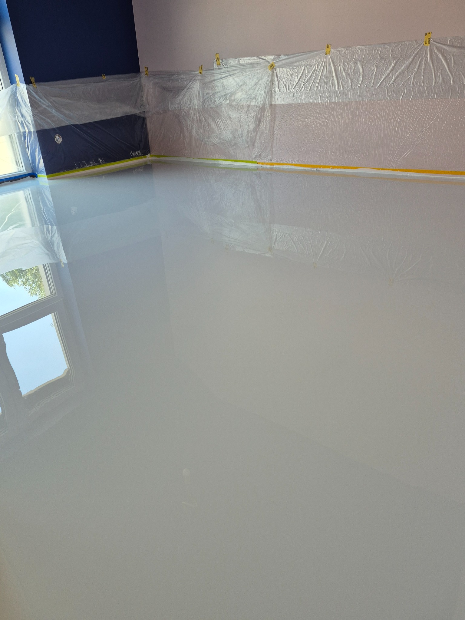 Błyszcząca, jasnoszara powierzchnia podłogi po aplikacji w Będzienicy. Ochronne folie przy ścianach i oknie. Widoczne odbicie okna w gładkiej powierzchni.