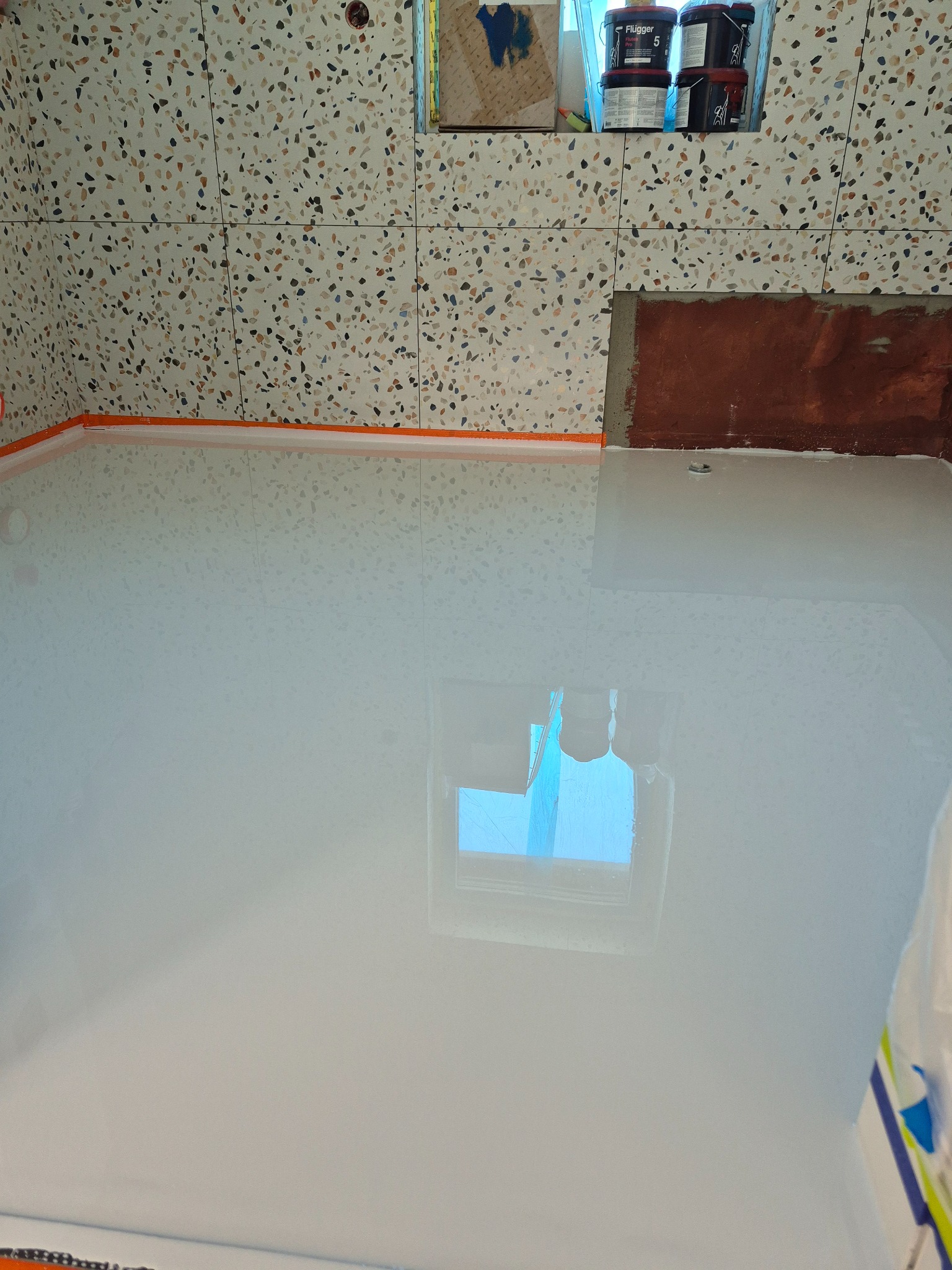 Biała, lśniąca podłoga po wylaniu, z widocznymi detalami wykończenia przy ścianach i terakotą w tle. Na ścianie puszki z farbą.