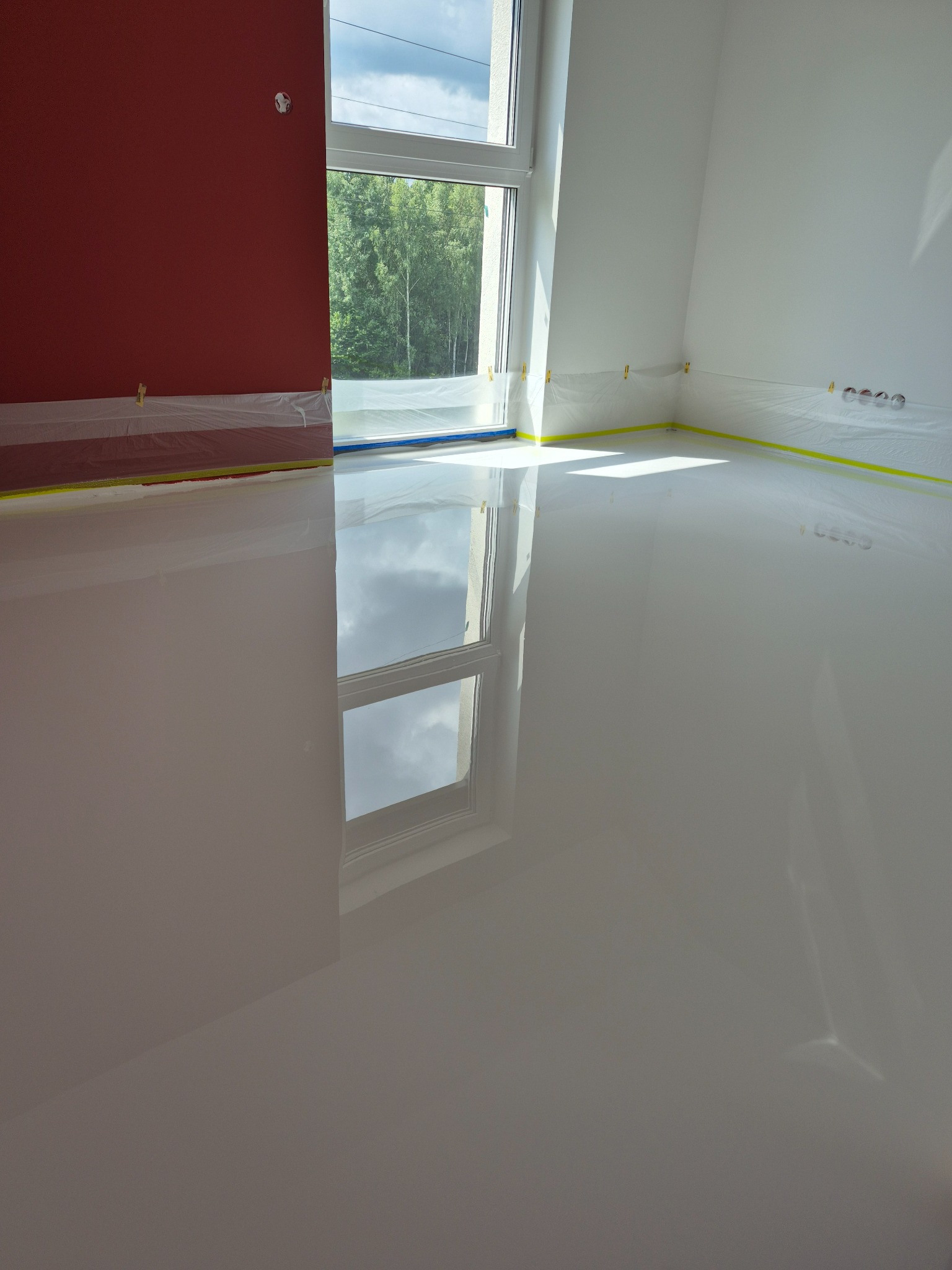 Biała, lśniąca podłoga w pokoju po wykonaniu wylewki. Okna zabezpieczone folią i taśmą malarską. Widoczne odbicie okna w podłodze.
